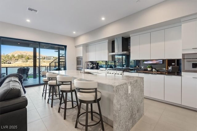 kitchen with wall chimney exhaust hood, a sink, a center island with sink, and white cabinetry