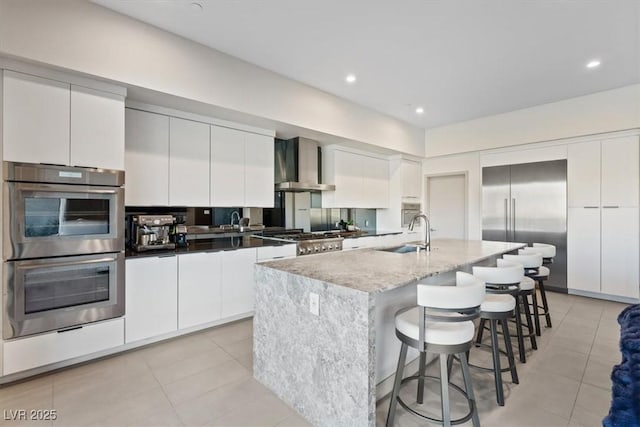 kitchen with stainless steel appliances, a sink, white cabinets, wall chimney range hood, and a center island with sink