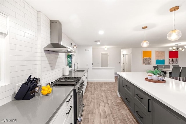 kitchen featuring light wood-type flooring, backsplash, wall chimney exhaust hood, stainless steel range with gas cooktop, and hanging light fixtures