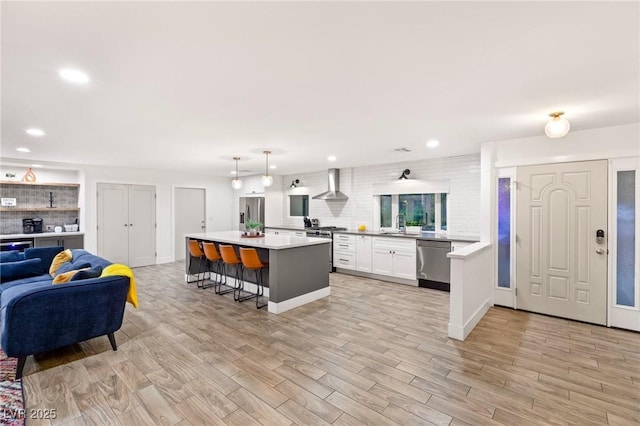 kitchen featuring wall chimney exhaust hood, stainless steel appliances, a kitchen breakfast bar, decorative light fixtures, and white cabinets