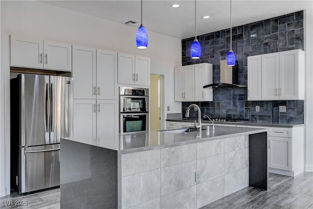 kitchen featuring pendant lighting, white cabinets, wall chimney range hood, an island with sink, and appliances with stainless steel finishes