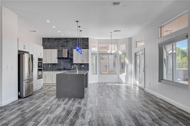 kitchen featuring stainless steel appliances, wall chimney range hood, pendant lighting, a center island with sink, and white cabinetry