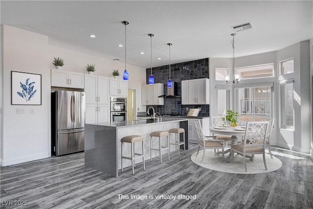 kitchen with hardwood / wood-style floors, a kitchen island with sink, wall chimney exhaust hood, appliances with stainless steel finishes, and white cabinetry