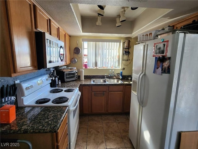 kitchen with sink, white appliances, light tile patterned flooring, a raised ceiling, and dark stone counters