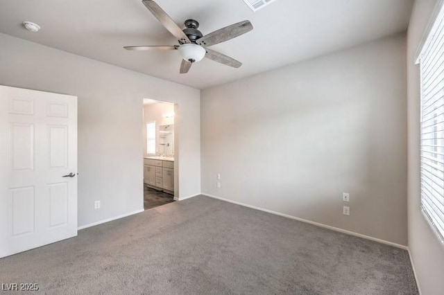 unfurnished bedroom featuring multiple windows, dark carpet, ensuite bath, and ceiling fan