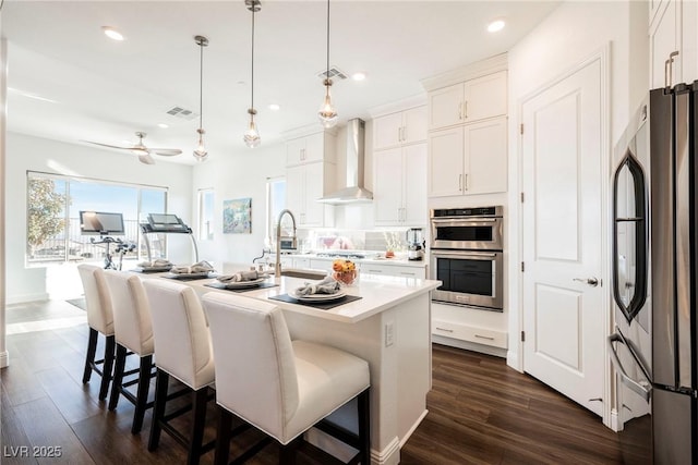 kitchen with a center island with sink, appliances with stainless steel finishes, hanging light fixtures, wall chimney exhaust hood, and white cabinets