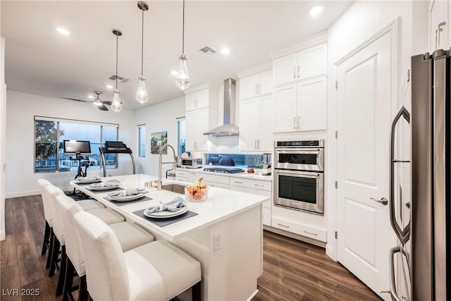 kitchen with a kitchen island with sink, a breakfast bar area, stainless steel appliances, white cabinetry, and wall chimney range hood