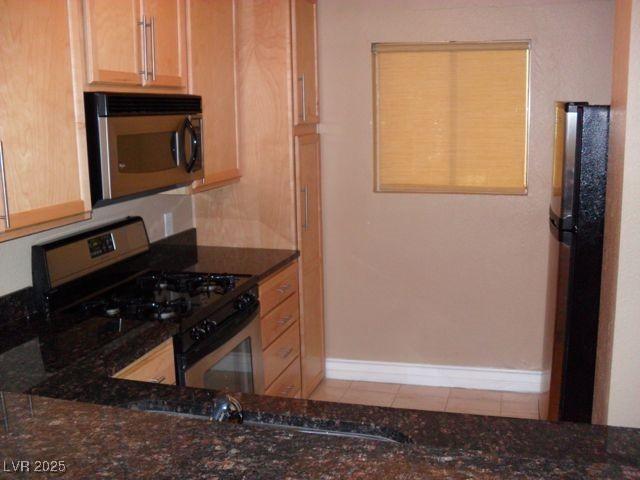 kitchen featuring black gas range, dark stone counters, refrigerator, light tile patterned floors, and light brown cabinetry