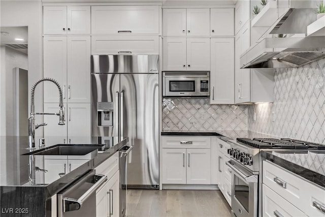 kitchen with white cabinets, wall chimney range hood, sink, light hardwood / wood-style flooring, and built in appliances