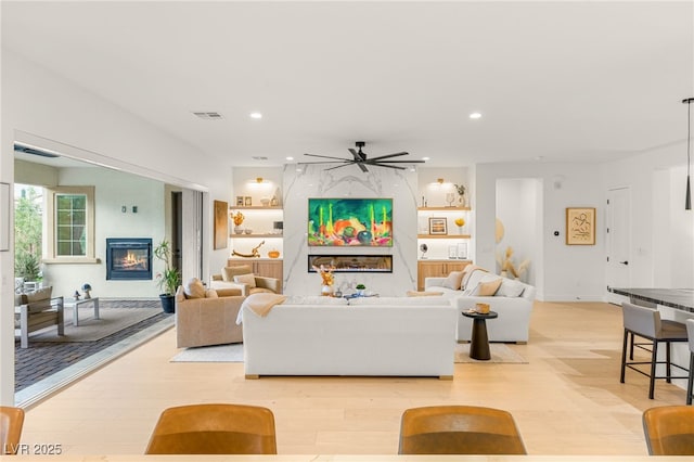 living room featuring built in shelves, ceiling fan, a fireplace, and light hardwood / wood-style floors