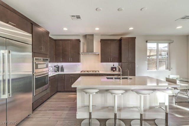 kitchen with a center island with sink, wall chimney exhaust hood, a kitchen bar, dark brown cabinetry, and stainless steel appliances