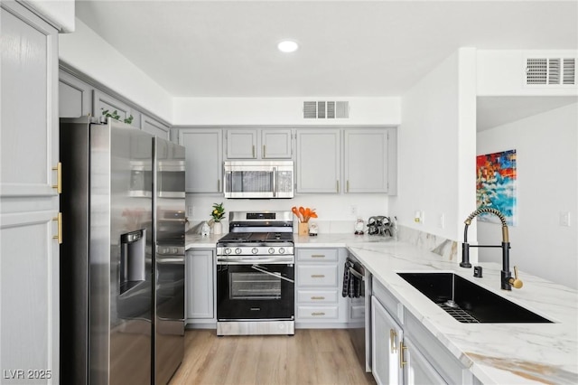 kitchen with gray cabinetry, light stone countertops, sink, stainless steel appliances, and light wood-type flooring