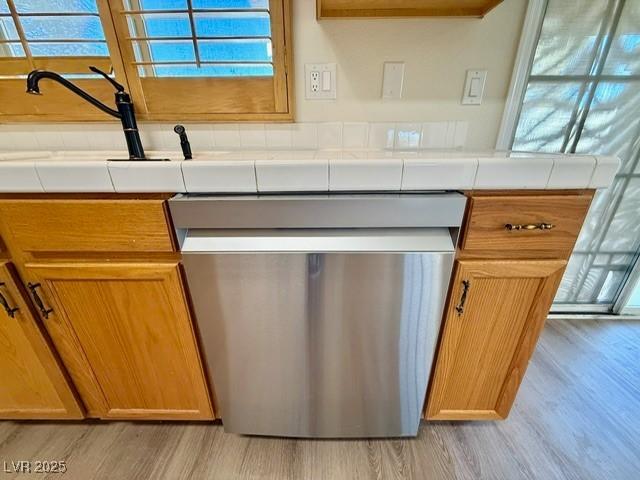 interior details featuring light wood-type flooring, tile countertops, and stainless steel dishwasher