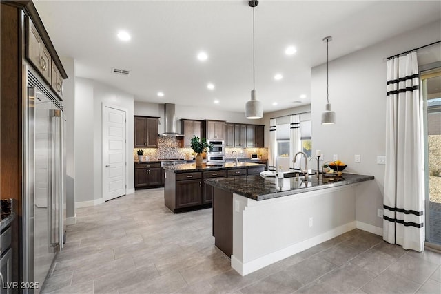 kitchen with stainless steel appliances, hanging light fixtures, wall chimney range hood, kitchen peninsula, and dark stone counters