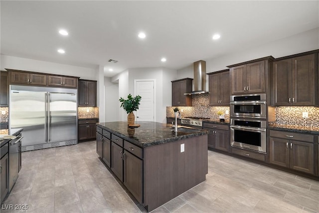 kitchen featuring wall chimney exhaust hood, dark stone countertops, dark brown cabinets, and appliances with stainless steel finishes