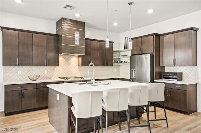 kitchen featuring wall chimney exhaust hood, dark brown cabinets, stainless steel appliances, sink, and an island with sink