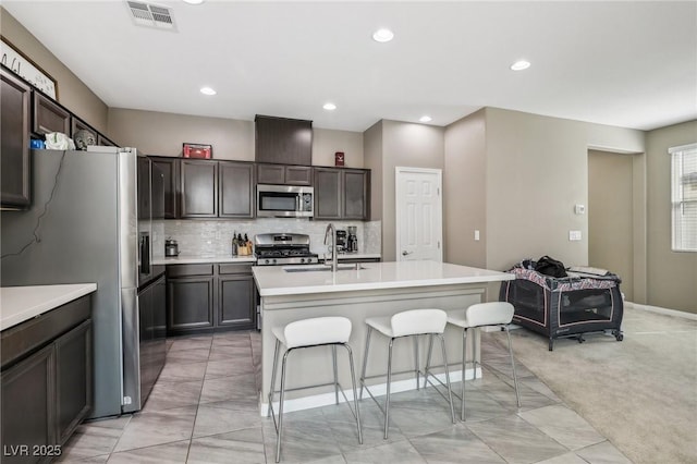 kitchen with a center island with sink, sink, appliances with stainless steel finishes, light colored carpet, and dark brown cabinetry