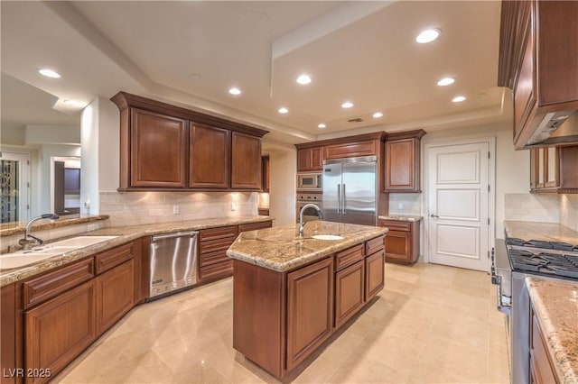 kitchen featuring a raised ceiling, built in appliances, light stone countertops, and a center island with sink