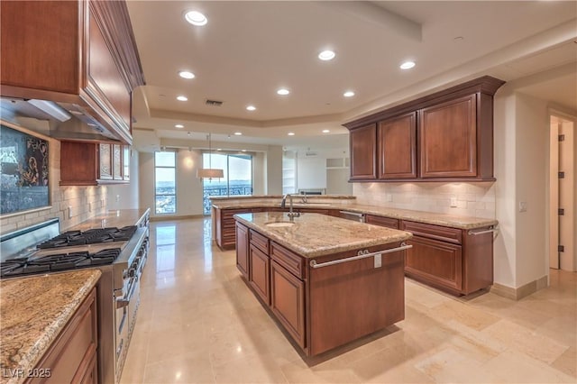 kitchen with sink, a raised ceiling, light stone counters, high end stove, and an island with sink