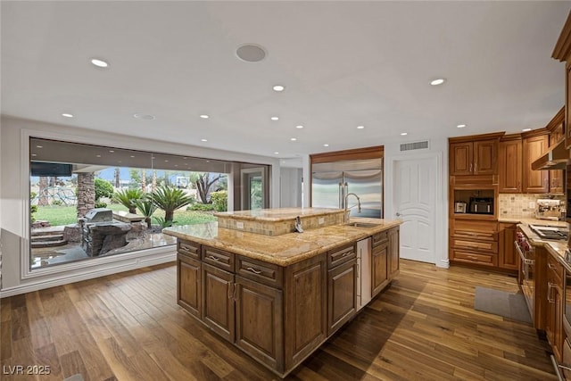 kitchen with a kitchen island with sink, dark wood-type flooring, stainless steel built in fridge, tasteful backsplash, and light stone counters