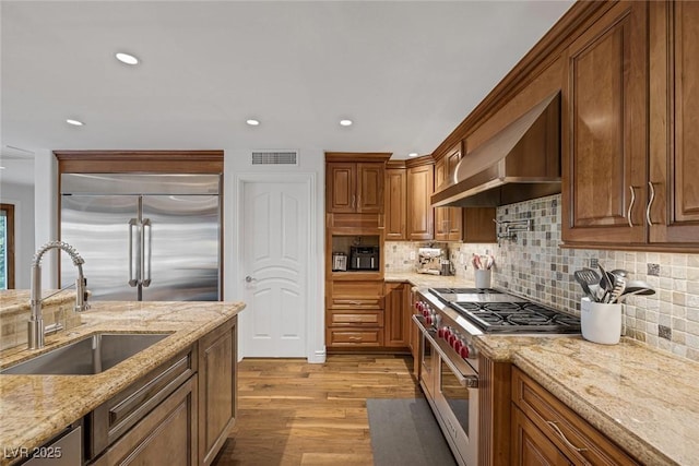 kitchen with light stone countertops, high end appliances, sink, wall chimney range hood, and light hardwood / wood-style flooring