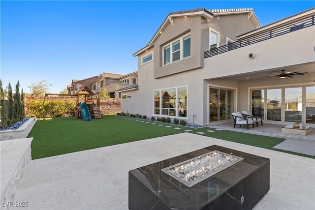 back of property featuring ceiling fan, a yard, an outdoor fire pit, a playground, and a patio area