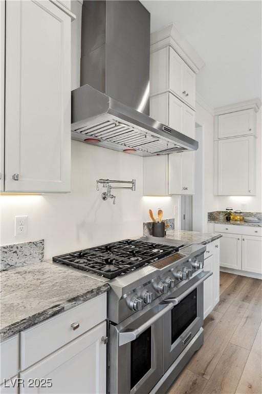 kitchen with double oven range, wall chimney range hood, light wood-type flooring, light stone counters, and white cabinetry