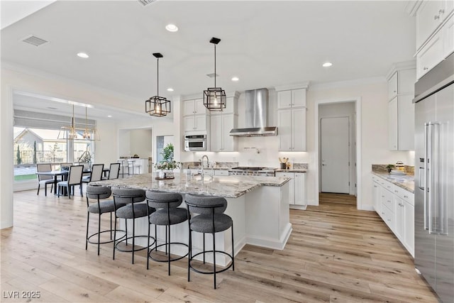 kitchen featuring stainless steel appliances, wall chimney range hood, an island with sink, decorative light fixtures, and white cabinets