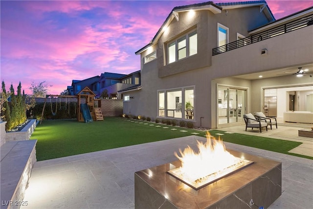 back house at dusk with a balcony, a fire pit, a patio area, a playground, and a lawn