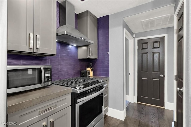 kitchen featuring tasteful backsplash, gray cabinetry, dark hardwood / wood-style flooring, stainless steel appliances, and wall chimney range hood