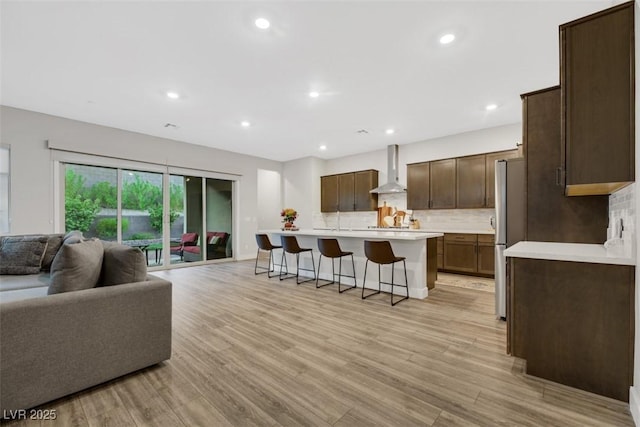 kitchen with decorative backsplash, wall chimney exhaust hood, a breakfast bar, a kitchen island with sink, and light hardwood / wood-style floors