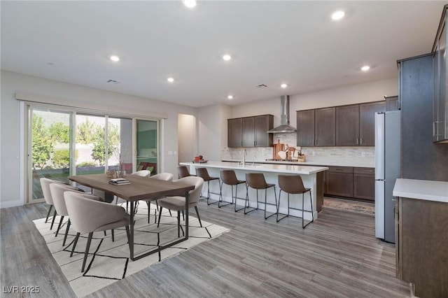 kitchen featuring a kitchen island with sink, wall chimney range hood, white fridge, light hardwood / wood-style floors, and decorative backsplash