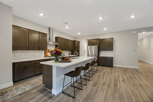 kitchen featuring a center island with sink, a kitchen breakfast bar, wall chimney range hood, sink, and stainless steel fridge