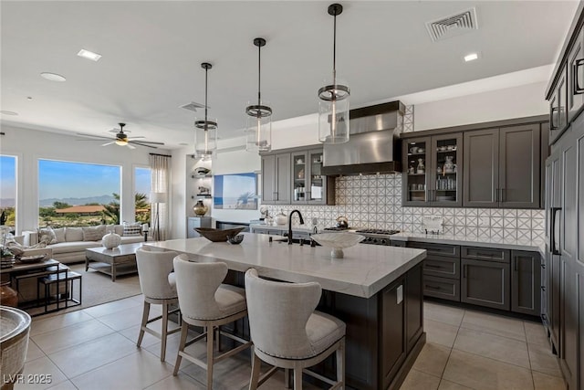 kitchen with sink, wall chimney exhaust hood, hanging light fixtures, an island with sink, and light tile patterned flooring