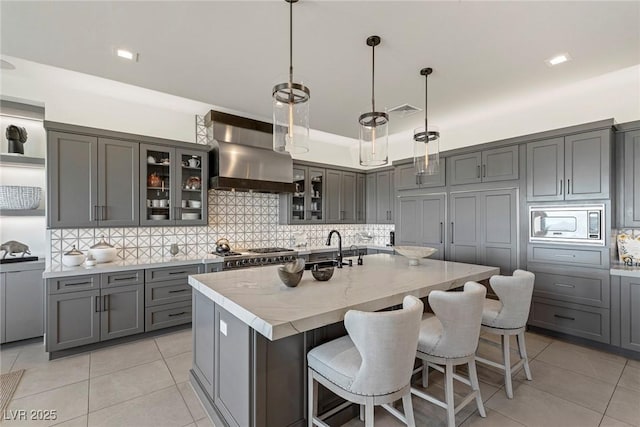 kitchen featuring pendant lighting, stainless steel microwave, a kitchen island with sink, wall chimney range hood, and gray cabinets