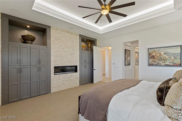 bedroom featuring ceiling fan, a stone fireplace, crown molding, a tray ceiling, and light carpet