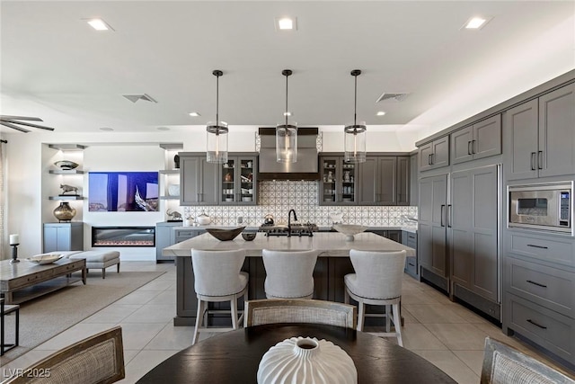 kitchen with stainless steel microwave, wall chimney exhaust hood, hanging light fixtures, an island with sink, and light tile patterned floors