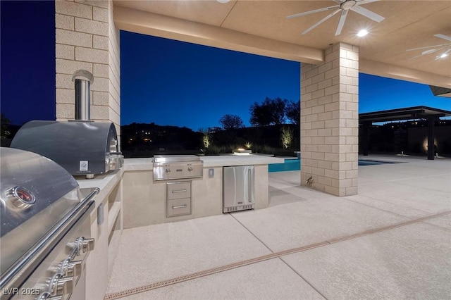 patio at night with grilling area, ceiling fan, and an outdoor kitchen