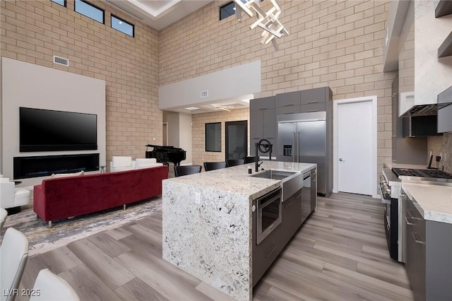 kitchen with a towering ceiling, sink, built in appliances, a center island with sink, and light hardwood / wood-style flooring