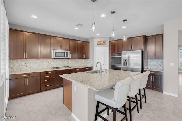 kitchen with light stone counters, a sink, appliances with stainless steel finishes, a center island with sink, and pendant lighting