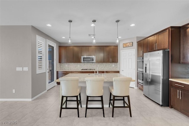 kitchen with appliances with stainless steel finishes, a center island with sink, hanging light fixtures, and light stone countertops