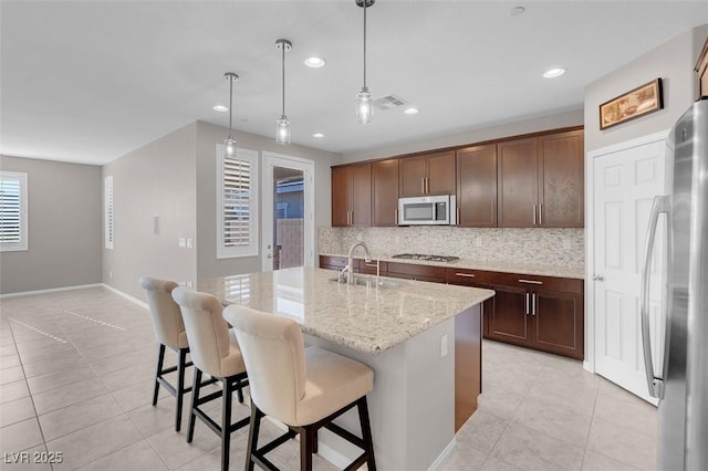 kitchen with light stone counters, a kitchen island with sink, stainless steel appliances, a sink, and pendant lighting