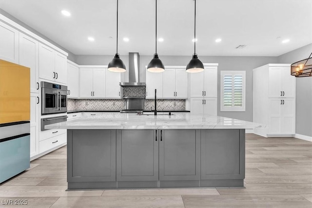 kitchen featuring an island with sink, wall chimney range hood, white cabinetry, and light stone countertops