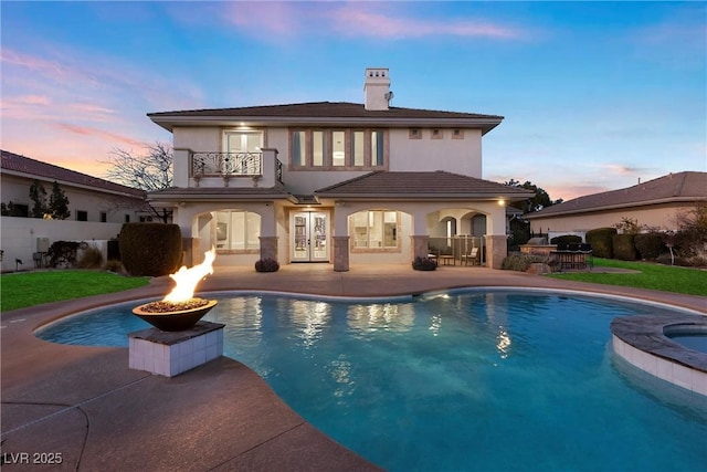 back house at dusk with a fire pit, a patio area, a balcony, and a swimming pool with hot tub