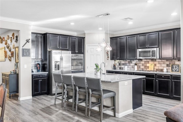 kitchen with a kitchen island with sink, hanging light fixtures, decorative backsplash, light stone countertops, and appliances with stainless steel finishes