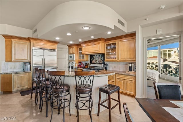 kitchen with built in appliances, light tile patterned floors, an island with sink, light stone countertops, and decorative backsplash