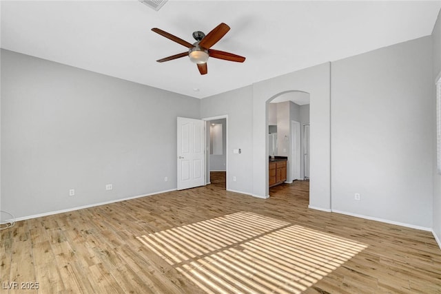 unfurnished bedroom featuring connected bathroom, baseboards, wood finished floors, arched walkways, and a ceiling fan