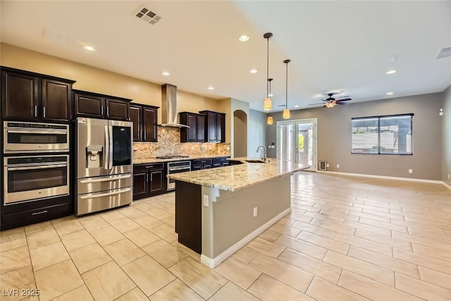 kitchen featuring a kitchen island with sink, wall chimney range hood, hanging light fixtures, sink, and appliances with stainless steel finishes