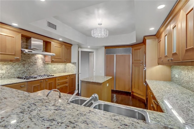 kitchen with paneled built in fridge, wall chimney exhaust hood, decorative backsplash, light stone counters, and stainless steel gas cooktop