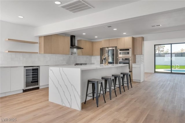 kitchen with wine cooler, a kitchen island with sink, wall chimney exhaust hood, and stainless steel appliances